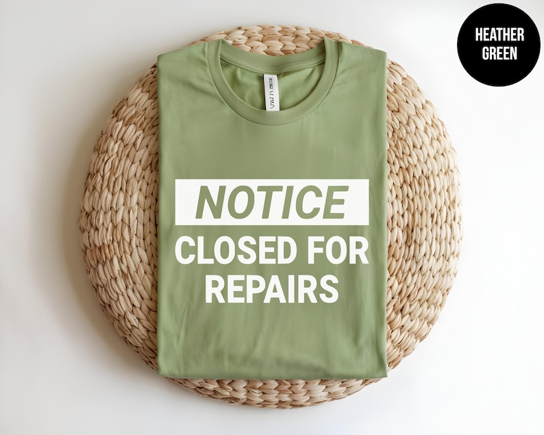 May include: A heather green t-shirt with the text "NOTICE CLOSED FOR REPAIRS" printed in white on a rectangular background.