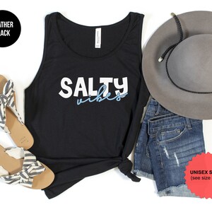 May include: Black tank top with the text "Salty Vibes" in white and blue lettering. The tank top is styled with denim shorts, a gray hat, and striped sandals.