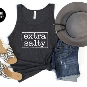 May include: A heather dark grey tank top with the text "extra salty" printed in white on the front. The tank top is styled with denim shorts, a grey fedora, and striped sandals.