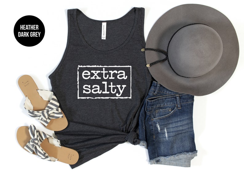 May include: A heather dark grey tank top with the text "extra salty" printed in white on the front. The tank top is styled with denim shorts, a grey fedora, and striped sandals.