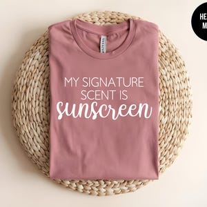 May include: A heather mauve t-shirt with the text "My Signature Scent Is Sunscreen" printed in white.