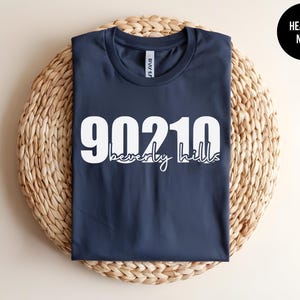 May include: A heather navy blue t-shirt featuring the white text 90210 in a large bold font with the words beverly hills written in a smaller white cursive script font underneath. A black circular badge in the corner displays the text HEATHER NAVY.