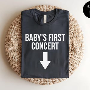 Pode incluir: Uma camiseta cinza escura mesclada com o texto "BABY'S FIRST CONCERT" e uma seta branca apontando para baixo.