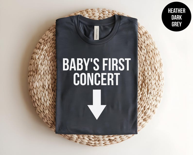 Pode incluir: Uma camiseta cinza escura mesclada com o texto "BABY'S FIRST CONCERT" e uma seta branca apontando para baixo.