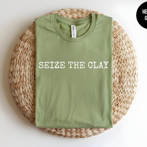 May include: A heather green t-shirt with a crew neckline and short sleeves. The shirt features the white text "SEIZE THE CLAY" printed in a bold, sans-serif font across the centre of the chest. A small black circle in the corner reads "HEATHER GREEN".
