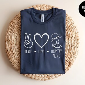 May include: A navy blue t-shirt with a white peace sign, heart, cowboy hat, and boots graphic. The text "Peace. Love. Country Music" is printed below the graphic.