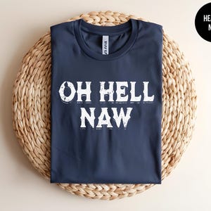 May include: A navy blue t-shirt featuring the phrase "OH HELL NAW" in distressed white text. The shirt is presented on a woven, circular mat. A black circle with "HEATHER NAVY" text is in the upper right corner.
