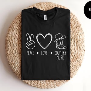 May include: Black t-shirt with a white peace sign, heart, and cowboy boots. The text "Peace, Love, Country Music" is printed below the symbols.