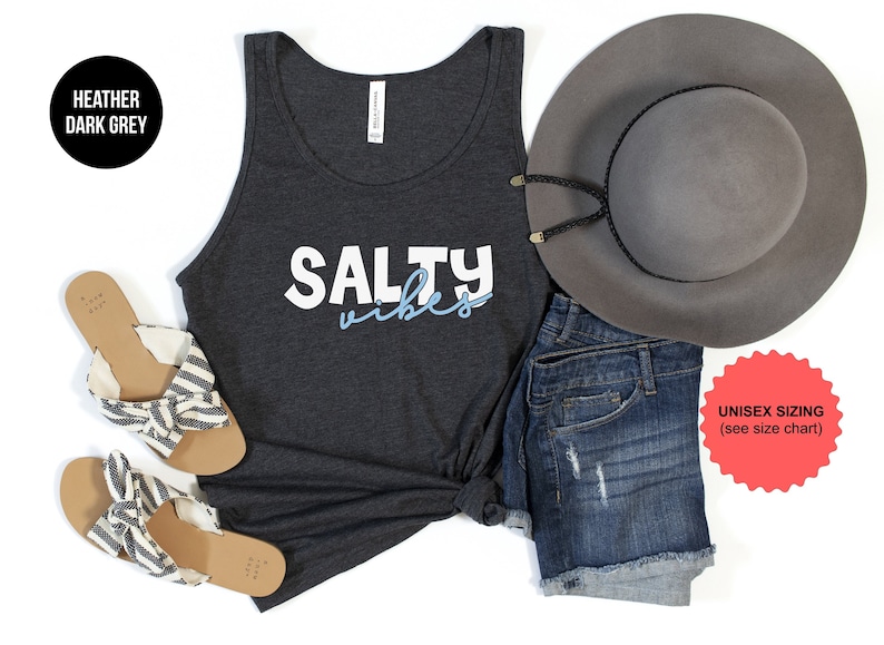 May include: A heather dark grey tank top with the text 'SALTY vibes' in white and blue lettering. The tank top is laying flat on a white surface with a pair of striped sandals, a pair of denim shorts, and a grey fedora hat. The text 'UNISEX SIZING (see size chart)' is on a red starburst sticker.