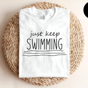 May include: White t-shirt with the text "just keep swimming" in black lettering. The shirt is on a woven straw background.