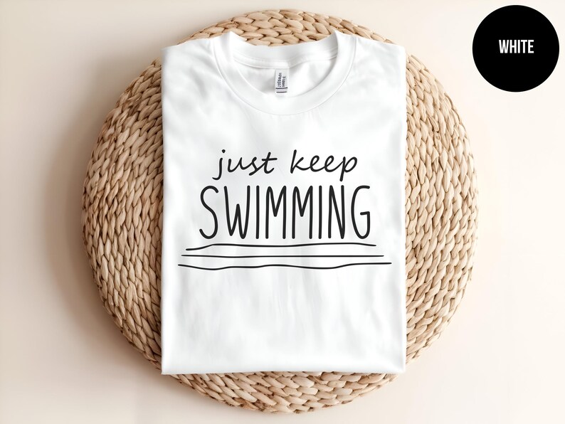 May include: White t-shirt with the text "just keep swimming" in black lettering. The shirt is on a woven straw background.