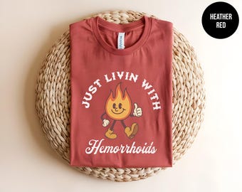 May include: Heather red t-shirt with the text "JUST LIVIN WITH Hemorrhoids" in white. The shirt features a cartoon flame character with a smiling face and thumbs up. The shirt is displayed on a woven surface.