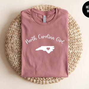 Pu&ograve; includere: T-shirt color malva melange con la scritta "North Carolina Girl" sopra un contorno bianco dello stato con un cuore. La maglietta &egrave; esposta su una superficie circolare intrecciata. Un cerchio nero con "Heather Mauve" &egrave; in alto a destra.
