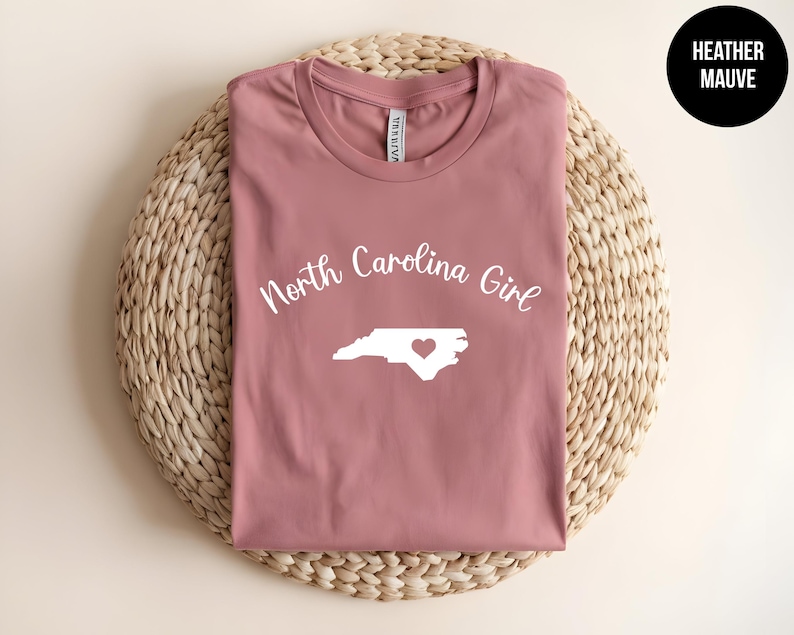 Pu&ograve; includere: T-shirt color malva melange con la scritta "North Carolina Girl" sopra un contorno bianco dello stato con un cuore. La maglietta &egrave; esposta su una superficie circolare intrecciata. Un cerchio nero con "Heather Mauve" &egrave; in alto a destra.