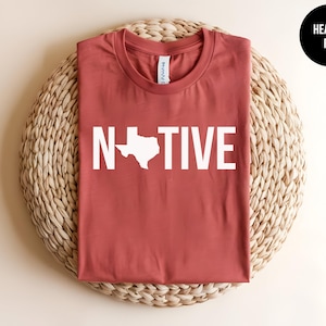 May include: A heather red t-shirt with the word "NATIVE" printed in white, featuring an outline of the state of Texas.