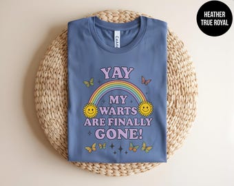 May include: A heather true royal blue t-shirt featuring a graphic with a rainbow, two yellow suns, and several butterflies. The text reads YAY MY WARTS ARE FINALLY GONE! in purple letters. The design includes small star accents surrounding the central text.
