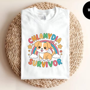 May include: White t-shirt featuring a cartoon dog with a rainbow, clouds, and flowers. The text on the shirt reads "CHLAMYDIA SURVIVOR" in colorful letters. The shirt is folded on a woven, circular surface.