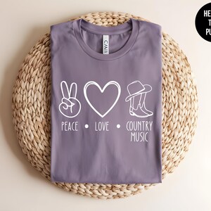 May include: Purple heather t-shirt with a white peace sign, heart, cowboy hat and boots, and the text "Peace Love Country Music".