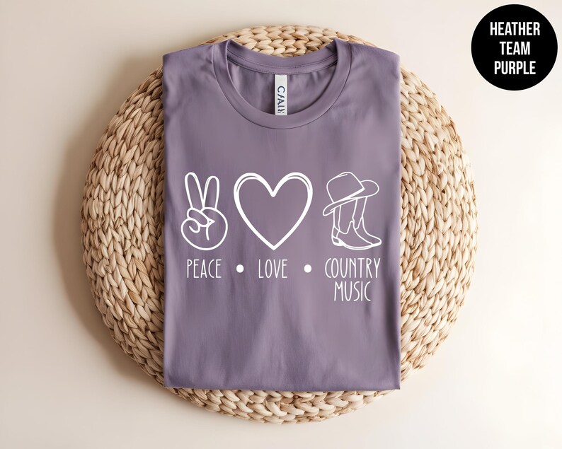 May include: Purple heather t-shirt with a white peace sign, heart, cowboy hat and boots, and the text "Peace Love Country Music".