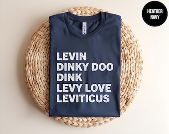 May include: A heather navy blue short-sleeved t-shirt with white distressed-style text printed on the front. The text reads: LEVIN, DINKY DOO, DINK, LEVY LOVE, LEVITICUS. The shirt features a crew neckline and a standard fit design.