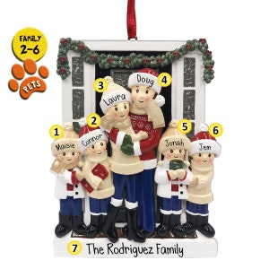 May include: A white Christmas ornament depicting a family of six standing in front of a house. The family is wearing winter clothing and holding gifts. The ornament is decorated with a red ribbon and the text "The Rodriguez Family".
