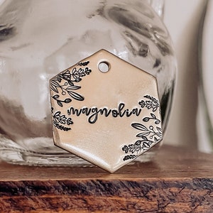 May include: A gold-toned metal tag with a hexagonal shape and a floral design. The tag has the word "magnolia" inscribed on it.