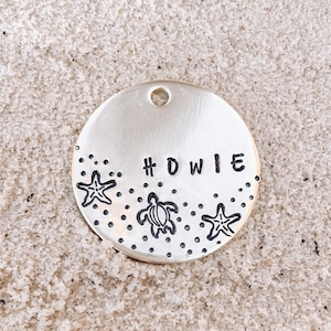May include: A round silver tag with the word "#DWIE" engraved on it. The tag is decorated with small dots and images of sea turtles and starfish.