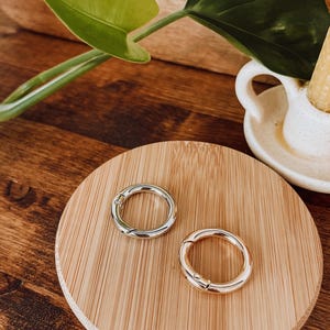 May include: Two metal round clasps, one silver and one gold, rest on a round bamboo coaster. The clasps are open and ready to be attached. The background includes a wooden surface and green plant leaves.