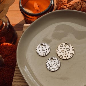 May include: Three circular silver-toned metal pet ID tags with engraved designs and names. One tag features the name Izzy with stars and flowers, one says Maple with coffee cups and pumpkins, and one says Chase with books and coffee beans.