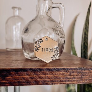 May include: A gold-toned hexagonal metal tag with a floral design and the name "LIDDY" engraved on it. The tag is hanging from a clear glass bottle with a cork stopper.