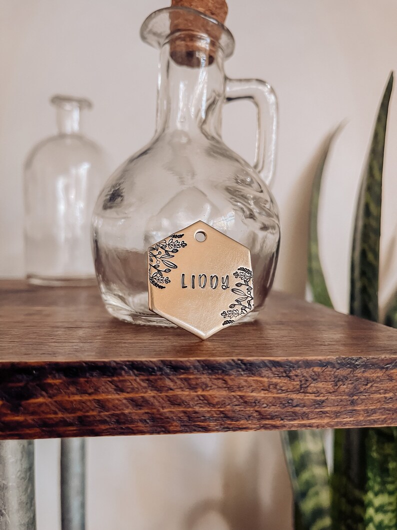 May include: A gold-toned hexagonal metal tag with a floral design and the name "LIDDY" engraved on it. The tag is hanging from a clear glass bottle with a cork stopper.