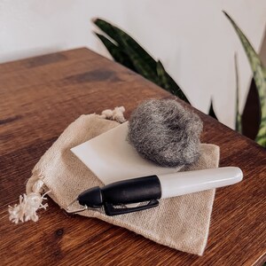 Pu&ograve; includere: Una piccola borsa di tessuto beige con un quadrato bianco e una palla di lana grigia all'interno. Un pennarello bianco e nero &egrave; appoggiato sulla borsa.