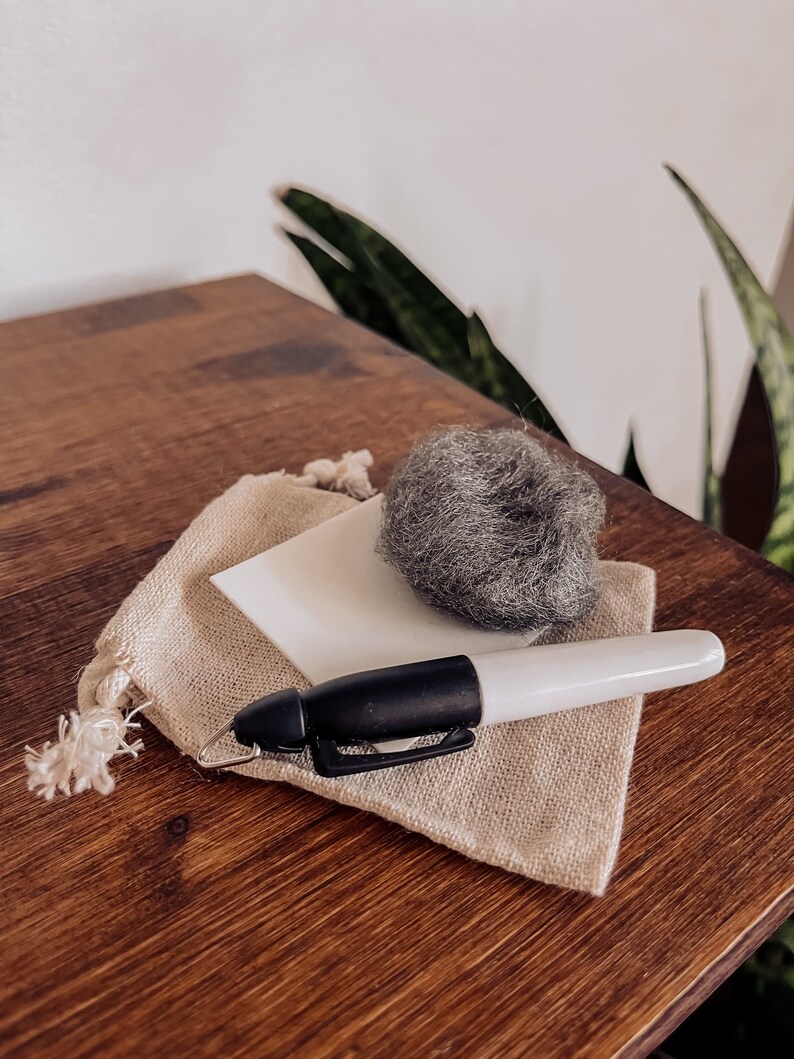 Pu&ograve; includere: Una piccola borsa di tessuto beige con un quadrato bianco e una palla di lana grigia all'interno. Un pennarello bianco e nero &egrave; appoggiato sulla borsa.