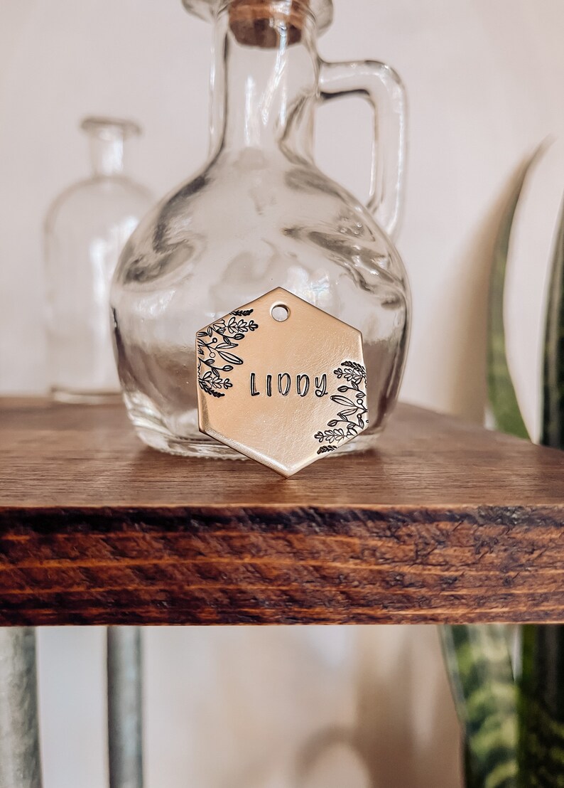 May include: A gold hexagonal metal tag with a floral design and the name "Liddy" engraved on it.
