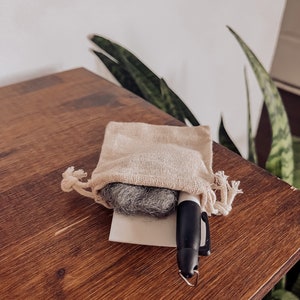 Pu&ograve; includere: Un piccolo panno per la pulizia grigio in una borsa beige con coulisse e un pennarello nero su una superficie di legno.
