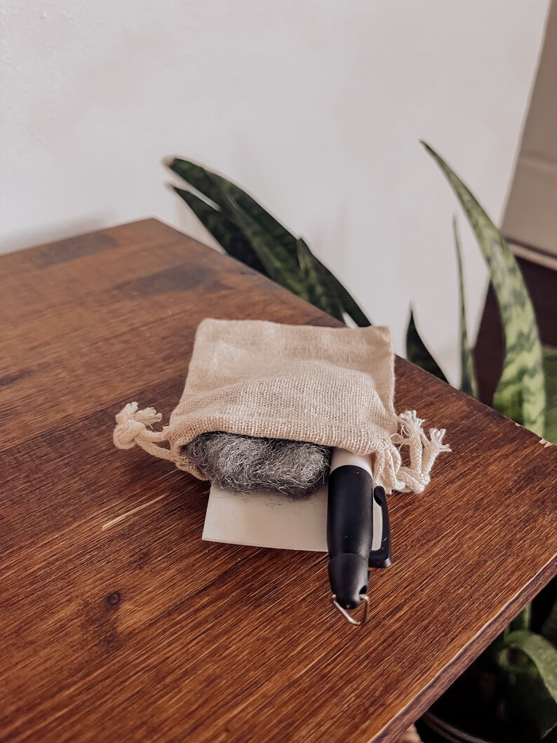 Pu&ograve; includere: Un piccolo panno per la pulizia grigio in una borsa beige con coulisse e un pennarello nero su una superficie di legno.