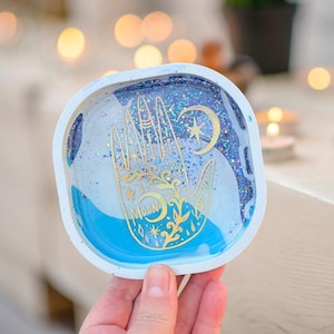 May include: A square, white-edged tray with a blue and glittery interior. The tray features a gold hand design with a crescent moon and star. The word "MOON" is also visible in gold. The tray is held by a hand.