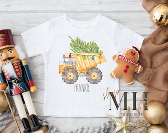 May include: White short-sleeved t-shirt featuring a watercolor illustration of a yellow dump truck carrying a Christmas tree, adorned with colorful lights. The name "FRANKIE" is printed below the truck. A gingerbread man and a nutcracker are also visible.
