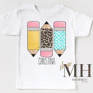 May include: White t-shirt with three colourful pencil graphics. The pencils have yellow, leopard print, and blue polka dot designs. The name "Christina" is printed below the pencils. The shirt also has a shop logo.