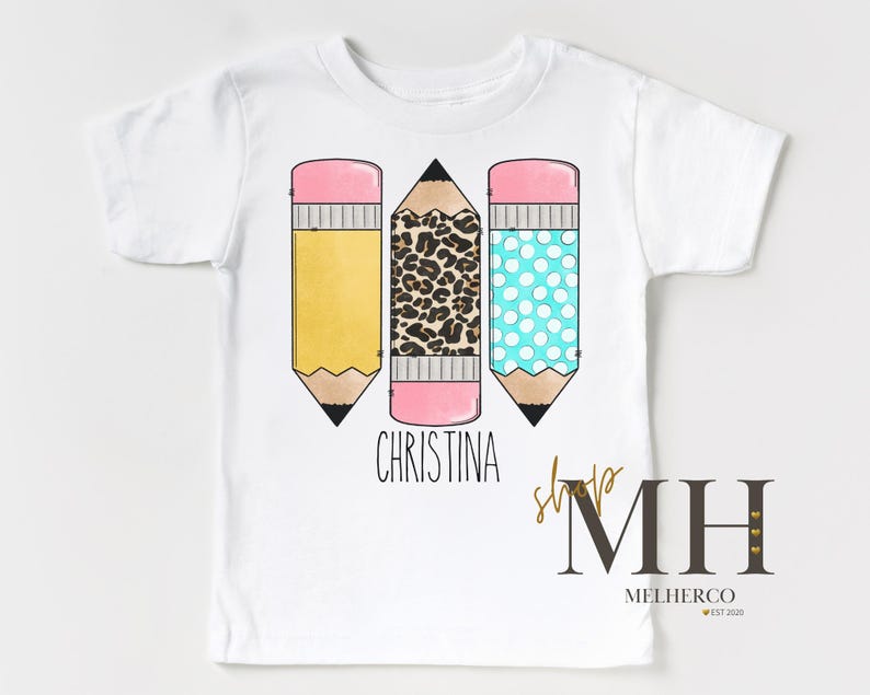 May include: White t-shirt with three colourful pencil graphics. The pencils have yellow, leopard print, and blue polka dot designs. The name "Christina" is printed below the pencils. The shirt also has a shop logo.