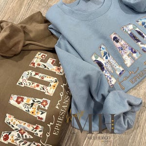 May include: Two sweatshirts, one brown and one light blue, are displayed. The word "MADE" is featured on each, with floral patterns within the letters. The text "for more EPHESIANS 2:10" is also visible. The sweatshirts are on a wooden surface.