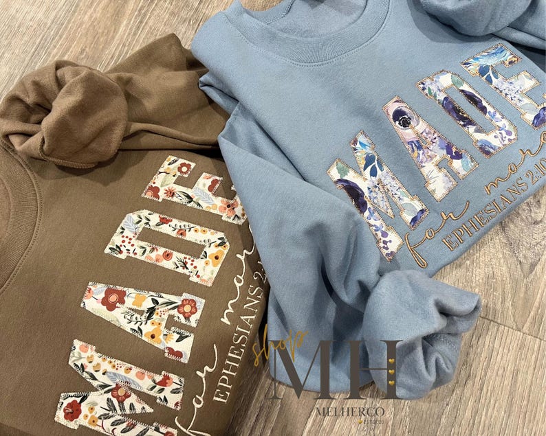 May include: Two sweatshirts, one brown and one light blue, are displayed. The word "MADE" is featured on each, with floral patterns within the letters. The text "for more EPHESIANS 2:10" is also visible. The sweatshirts are on a wooden surface.