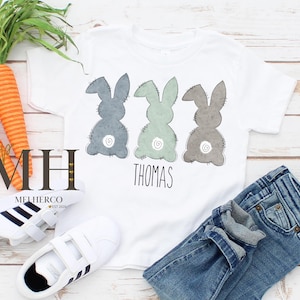 May include: White t-shirt featuring three bunny silhouettes in blue, green, and gray, with the name "Thomas" below. The shirt is paired with blue jeans and white sneakers. Carrots are also in the image.