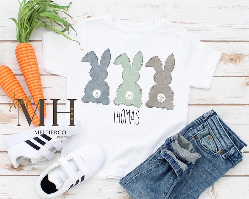 May include: White t-shirt featuring three bunny silhouettes in blue, green, and gray, with the name "Thomas" below. The shirt is paired with blue jeans and white sneakers. Carrots are also in the image.