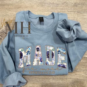 May include: A light blue sweatshirt with the word "MADE" in large, floral-patterned letters. The words "for more" and "Ephesians 2:10" are embroidered in gold script below the main text. The sweatshirt is folded on a wooden surface.