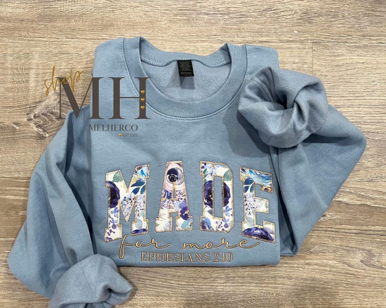 May include: A light blue sweatshirt with the word "MADE" in large, floral-patterned letters. The words "for more" and "Ephesians 2:10" are embroidered in gold script below the main text. The sweatshirt is folded on a wooden surface.