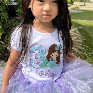 May include: A white t-shirt with a mermaid and the number 3 embroidered on it. The shirt also has the name "Miya" embroidered on it. The girl is wearing a purple and green tulle tutu.