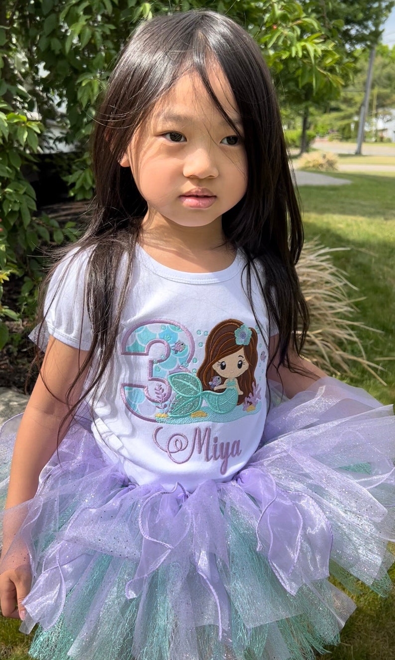 May include: A white t-shirt with a mermaid and the number 3 embroidered on it. The shirt also has the name "Miya" embroidered on it. The girl is wearing a purple and green tulle tutu.