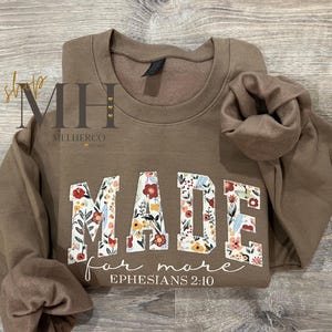 May include: A brown sweatshirt with the words "MADE for more" in floral print letters. The text "Ephesians 2:10" is written below the main text. The sweatshirt is folded on a wooden surface.
