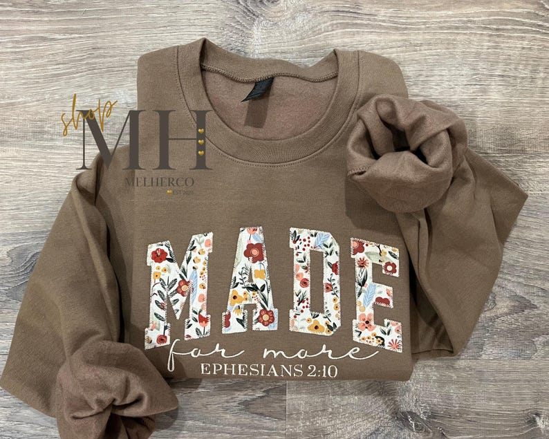 May include: A brown sweatshirt with the words "MADE for more" in floral print letters. The text "Ephesians 2:10" is written below the main text. The sweatshirt is folded on a wooden surface.